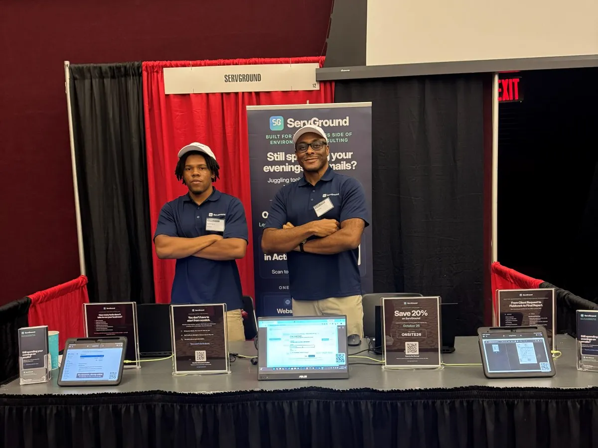 David Obim and a colleague in ServGround shirts and caps standing at the ServGround booth, with the app running on screens on the table
